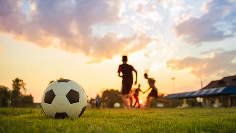 Im Vordergrund ein Fußball auf einer Wiese, im Hintergrund zwei Silhoutten im Sonnenuntergang
