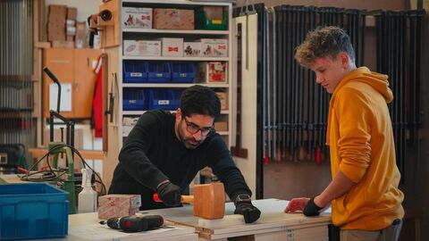 Wirtschaftsminister Mansoori steht in einer Werkstatt über einen Tisch gebeugt mit einem Hammer. Neben ihm steht ein junger Mann, der seine Tätigkeit beobachtet.