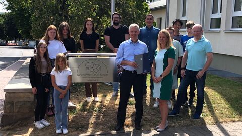 Staatssekretär Lösel besucht Lerncamp der Geschwister-Scholl-Schule Alsfeld. Er steht gemeinsam mit Schülerinnen und Schülern sowie Lehrerinnen und Lehrern vor dem Schild der Schule.